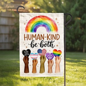 Könnte beinhalten: Eine weiße Gartenflagge mit einem Regenbogen und den Worten "HUMAN-KIND be both". Die Flagge zeigt Hände, die herzförmige Symbole halten, die Black Lives Matter, Transgender-Stolz, die amerikanische Flagge, Frieden und LGBTQ+-Stolz repräsentieren.