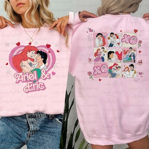May include: Pink sweatshirt featuring a graphic of Ariel and Eric embracing within a heart, with the text "Ariel & Eric." The back of the sweatshirt displays a grid of Ariel and Eric illustrations. Hearts and floral accents complete the design.