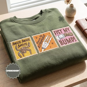 May include: An olive green crewneck sweatshirt featuring three rectangular embroidered patches designed like postage stamps. The patches display a brown creature with the text "AMAZE AMAZE AMAZE," a rocket with "Power by ASTROPHAGE," and a fist bump with "FIST MY BUMP!"