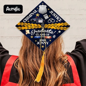 May include: A navy blue graduation cap with a Criminal Justice theme. The cap features a police badge, handcuffs, and a police car graphic. Text includes "Criminal Justice", "Graduate Class of 2026", and the name "Jonny". A yellow tassel hangs from the cap.