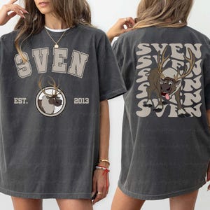 Pode incluir: Camiseta cinza escuro com o nome "SVEN" em um design de letras em bloco na frente e nas costas. A frente apresenta um gr&aacute;fico de rena e o texto "EST. 2013". As costas t&ecirc;m um gr&aacute;fico de rena e o nome "SVEN" repetido.