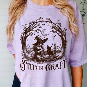 May include: A light purple t-shirt featuring a dark brown graphic of a witch in a pointed hat knitting with a black cat nearby. The scene is framed by bare tree branches, a crescent moon, and bats. The text STITCH CRAFT is printed below.