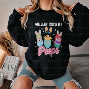 May include: Black sweatshirt with the text "CHILLIN' WITH MY Peeps". The graphic features three cartoon characters in Easter egg designs. The sweatshirt is a casual style.
