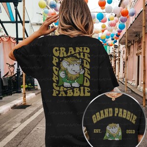May include: Black t-shirt with a graphic print of a cartoon character with the text "Grand Pabbie" in yellow letters. The character is wearing a green robe and has a white beard. The text "Grand Pabbie" is also printed on a black t-shirt with a white border.