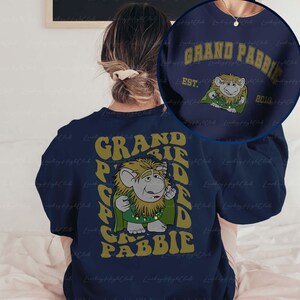 May include: A navy blue sweatshirt with a graphic of a green and yellow creature with the text "Grand Pabbie" in yellow letters. The text is repeated in a vertical pattern on the back of the sweatshirt.