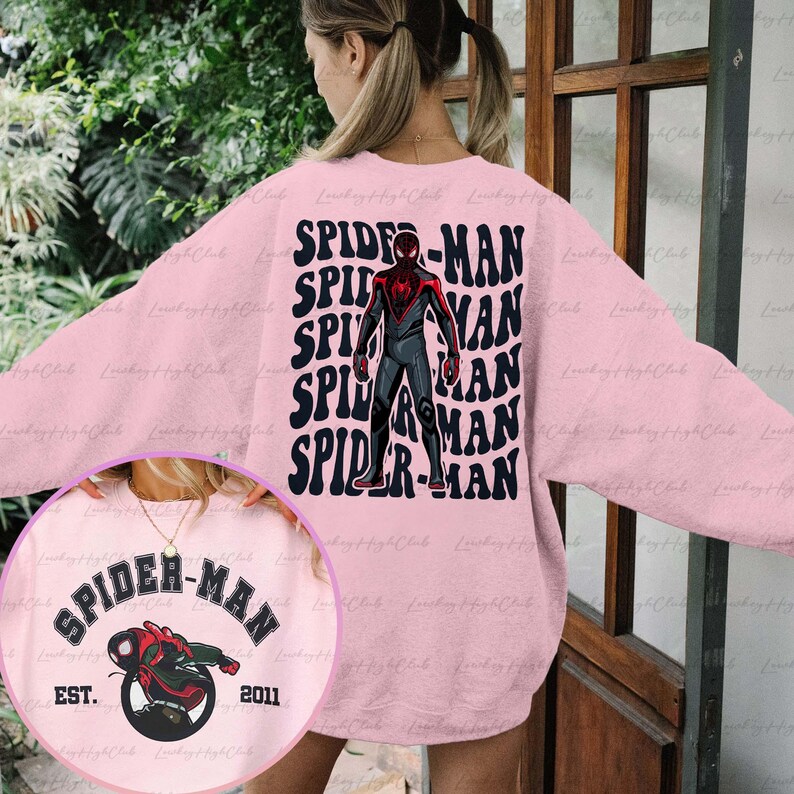 May include: Pink sweatshirt featuring a graphic of Spider-Man in a red and black suit. The text "SPIDER-MAN" is repeated in a retro font on the sweatshirt. A smaller graphic on the front of the sweatshirt shows Spider-Man in a circle with the text "SPIDER-MAN EST. 2011".