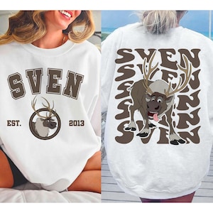 Pode incluir: Camisola branca com o nome "SVEN" em letras castanhas e um gr&aacute;fico de rena. O design frontal inclui "EST. 2013". A parte de tr&aacute;s apresenta um gr&aacute;fico de rena maior e o nome "SVEN" repetido num padr&atilde;o ondulado.
