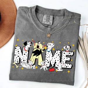 May include: Gray Comfort Colors t-shirt with a graphic featuring Dalmatian puppies and the word "NAME" in a dotted font. The design includes a cartoon villain and yellow star accents. A silver necklace is draped across the shirt.