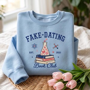 May include: A light blue crewneck sweatshirt featuring a graphic print with the text FAKE-DATING Book Club EST. 2021. The design includes a laboratory flask filled with pink hearts resting on a stack of three books, flanked by science icons.