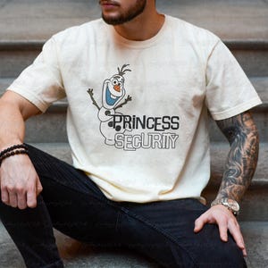 以下が含まれることがあります： クリーム色のTシャツ。漫画のキャラクター、オラフと黒字で「PRINCESS SECURITY」の文字がプリントされています。デザインにはオラフの笑顔と腕が含まれています。このTシャツはカジュアルウェアです。