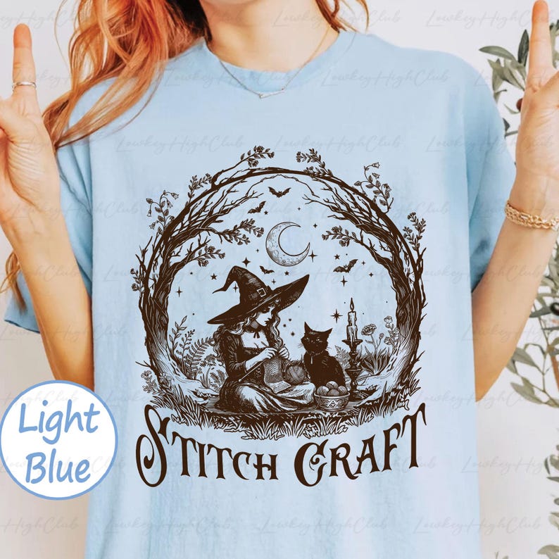 May include: A light blue t-shirt featuring a dark brown graphic of a witch wearing a pointed hat and knitting, accompanied by a black cat, a crescent moon, bats, and foliage. The text STITCH CRAFT is printed in a decorative font below the illustration.