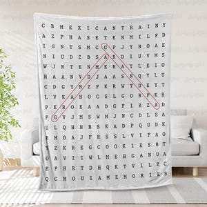 May include: White blanket with a word search puzzle design. The puzzle features a grid of letters with the words "MEXICAN," "TRAINY," and "MEMORIES" visible. A red line highlights a word. The blanket is draped over a white sofa.