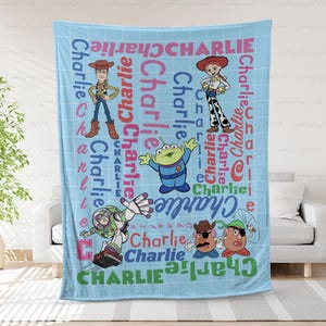 May include: A light blue fleece blanket featuring a white grid pattern and the name Charlie repeated in various colors and fonts. The design includes Toy Story characters Woody, Jessie, an alien, Buzz Lightyear, and Mr. Potato Head.