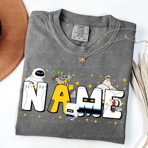 Può includere: T-shirt grigia Comfort Colors con la scritta "NAME" in un font a cartoni animati, con personaggi del film Wall-E. La maglietta ha uno scollo tondo e una piccola etichetta sulla parte superiore della schiena.
