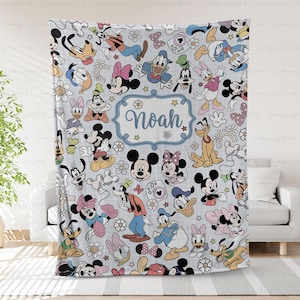 Pode incluir: Um cobertor cinza claro com um padrão de personagens de desenhos animados, incluindo Mickey Mouse, Minnie Mouse, Pato Donald e Pateta. O nome "Noah" é exibido em uma moldura azul. O cobertor está sobre um sofá branco.