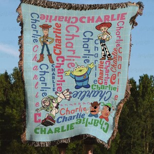 May include: A light blue woven throw blanket featuring a grid pattern and the name Charlie repeated in various colors and orientations. The blanket displays cartoon characters including Woody, Jessie, an alien, Buzz Lightyear, and Mr. Potato Head.