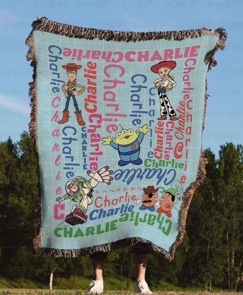 May include: A light blue woven throw blanket featuring a grid pattern and the name Charlie repeated in various colors and orientations. The blanket displays cartoon characters including Woody, Jessie, an alien, Buzz Lightyear, and Mr. Potato Head.