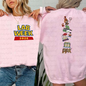 May include: A light pink sweatshirt with the text "LAB WEEK 2026" in yellow and blue, with a red banner. The back of the sweatshirt features various science-themed graphics in a vertical line, including a microscope and beakers.