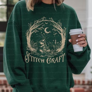May include: A dark green crewneck sweatshirt featuring a beige graphic print of a witch knitting with a black cat. The design is framed by tree branches, a crescent moon, and stars, with the text STITCH CRAFT printed in a decorative font below.