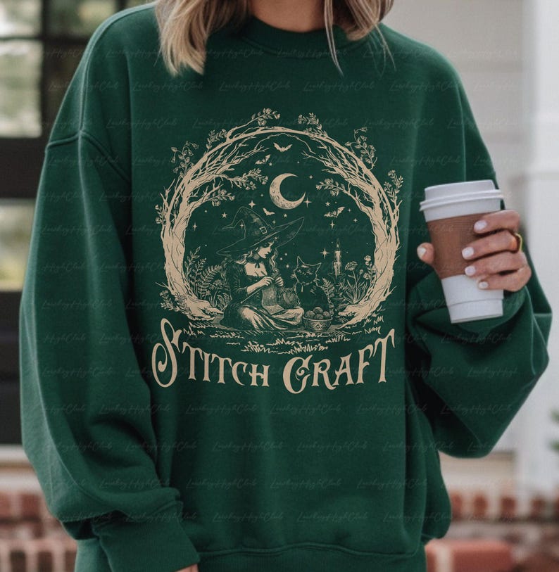 May include: A dark green crewneck sweatshirt featuring a beige graphic print of a witch knitting with a black cat. The design is framed by tree branches, a crescent moon, and stars, with the text STITCH CRAFT printed in a decorative font below.