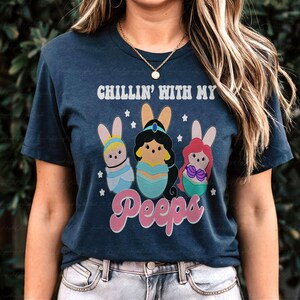 May include: Navy blue t-shirt with the text "CHILLIN' WITH MY Peeps". The shirt features a graphic of three cartoon characters dressed as Easter bunnies. The characters are in pastel colors with bunny ears.