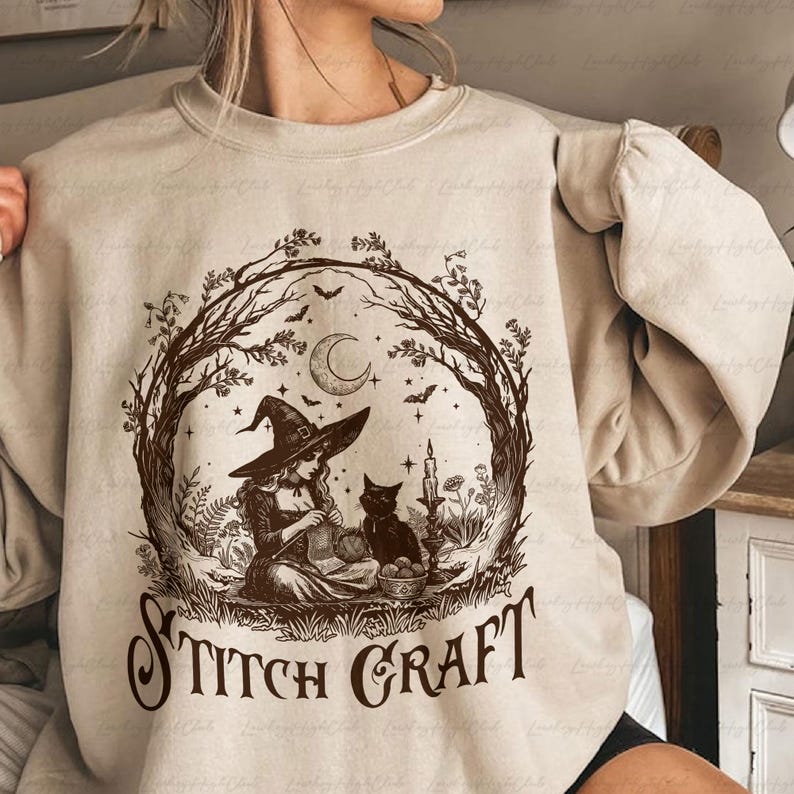 May include: A beige crewneck sweatshirt featuring a dark brown graphic of a witch wearing a pointed hat, knitting while sitting next to a black cat. The scene is framed by bare tree branches, a crescent moon, and bats, with the text STITCH CRAFT.