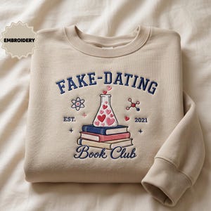 May include: A beige crewneck sweatshirt featuring dark blue embroidered text that reads FAKE-DATING BOOK CLUB. The design includes a stack of three books, a laboratory flask filled with pink and red hearts, an atom, and the text EST. 2021.