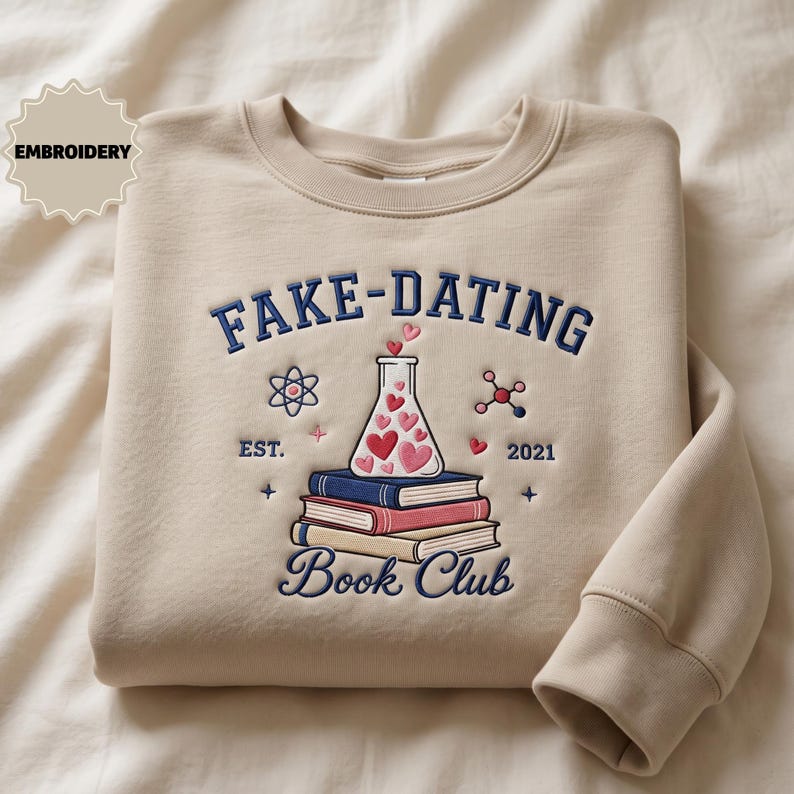 May include: A beige crewneck sweatshirt featuring dark blue embroidered text that reads FAKE-DATING BOOK CLUB. The design includes a stack of three books, a laboratory flask filled with pink and red hearts, an atom, and the text EST. 2021.