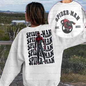May include: White sweatshirt with a black and red Spider-Man graphic on the front. The graphic is repeated multiple times in a stacked pattern. The text "SPIDER-MAN" is written in black with a white outline. The text "EST. 2011" is written in black below the graphic.