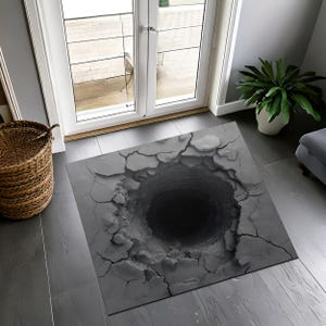 Könnte beinhalten: Grauer rechteckiger Teppich mit einem 3D-Design eines tiefen Lochs. Die Oberfläche des Teppichs ist um das Loch herum gerissen und erzeugt eine realistische Illusion. Der Teppich liegt auf einem dunkelgrauen Holzboden.
