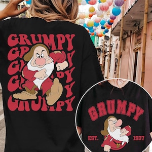 Peut inclure: T-shirt noir avec un motif rétro rouge représentant le mot "GRUMPY" et une illustration de Grumpy de Blanche-Neige. Le design comprend une image de Grumpy en tenue rouge. Une petite image du même motif est visible.