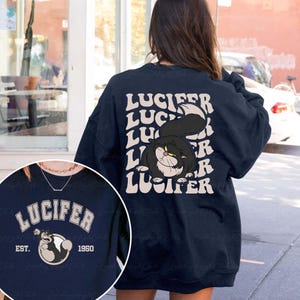 May include: Navy blue sweatshirt featuring a cartoon image of a black cat with the name "Lucifer" in white text. The design is repeated on the back. The sweatshirt also has a smaller design with the word "Lucifer" and the year "1950".