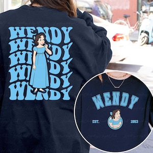 Puede incluir: Sudadera azul marino con el nombre "WENDY" en azul claro, y una ilustración de dibujos animados de una mujer con un vestido azul. El diseño se repite en la parte posterior. Un diseño circular en la parte delantera incluye el nombre "WENDY" y el año "1953".