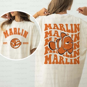 May include: Cream-colored t-shirt with orange text and graphics. The front features the word "MARLIN" above a cartoon basketball and the text "EST. 2003". The back displays the word "MARLIN" repeated with a cartoon fish.