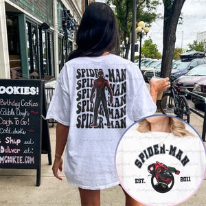 May include: White t-shirt with a black and red Spider-Man graphic on the back. The graphic features the text "SPIDER-MAN" repeated in a retro style. The front of the shirt features a smaller Spider-Man graphic with the text "SPIDER-MAN EST. 2011".