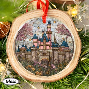 May include: A round glass ornament with a stained-glass design of a pink and blue castle, set within a wooden frame. The castle is surrounded by flowers and trees, with a red ribbon for hanging. The word "Glass" is printed on the bottom left.