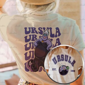 Pu&ograve; includere: Due magliette bianche con una grafica viola e nera di Ursula de La Sirenetta. La prima maglietta ha il testo "URSULA" ripetuto in un font retr&ograve;. La seconda maglietta ha il testo "URSULA" in un font universitario con il testo "EST. 1989" sotto.