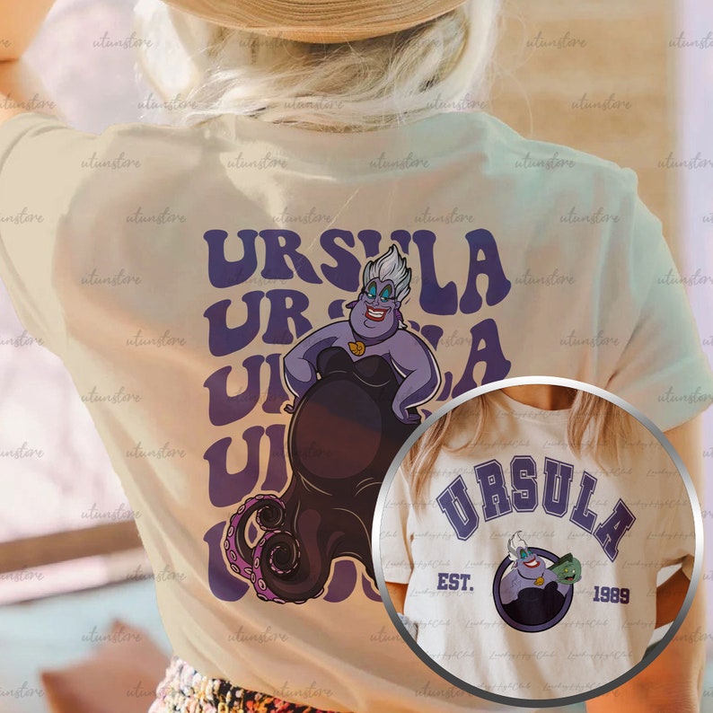 Pu&ograve; includere: Due magliette bianche con una grafica viola e nera di Ursula de La Sirenetta. La prima maglietta ha il testo "URSULA" ripetuto in un font retr&ograve;. La seconda maglietta ha il testo "URSULA" in un font universitario con il testo "EST. 1989" sotto.