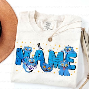 May include: Cream-colored t-shirt with a blue cat design spelling out "NAME". The design includes cartoon cats, stars, and a bird wearing a hat. A gold necklace and a brown hat are visible.