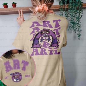 May include: A beige t-shirt with a purple monster graphic and the text "ART ART" printed on the back. The front of the shirt has a purple graphic with the text "ART EST. 2013".
