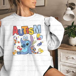 Op de afbeelding: Wit sweatshirt met een blauw Stitch-personage uit Lilo & Stitch, omringd door puzzelstukjes, vogels en een lint. De tekst "AUTISM" staat in regenboogkleuren boven het personage.