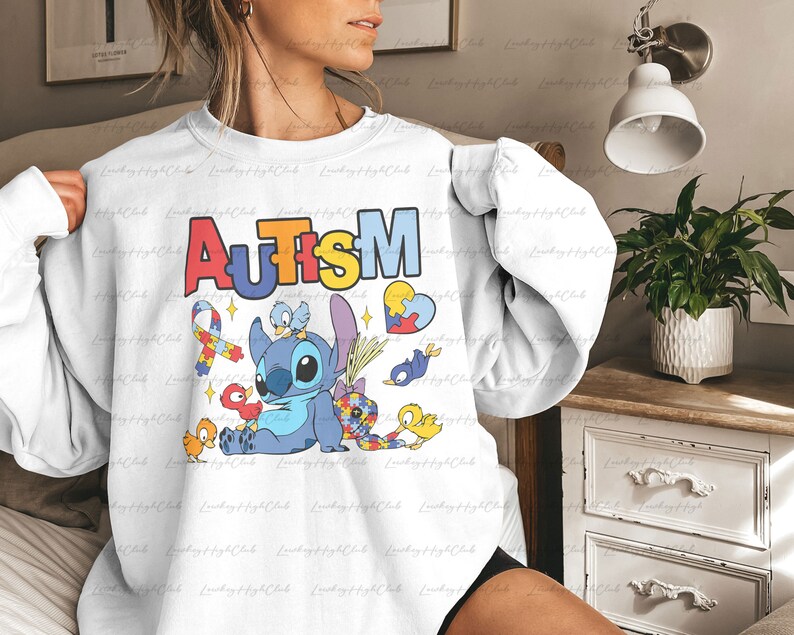 Op de afbeelding: Wit sweatshirt met een blauw Stitch-personage uit Lilo & Stitch, omringd door puzzelstukjes, vogels en een lint. De tekst "AUTISM" staat in regenboogkleuren boven het personage.