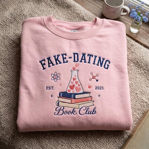 May include: A light pink crewneck sweatshirt featuring dark blue embroidered text that reads FAKE-DATING BOOK CLUB. The design includes a central Erlenmeyer flask filled with pink hearts, resting on a stack of three books, with science icons.