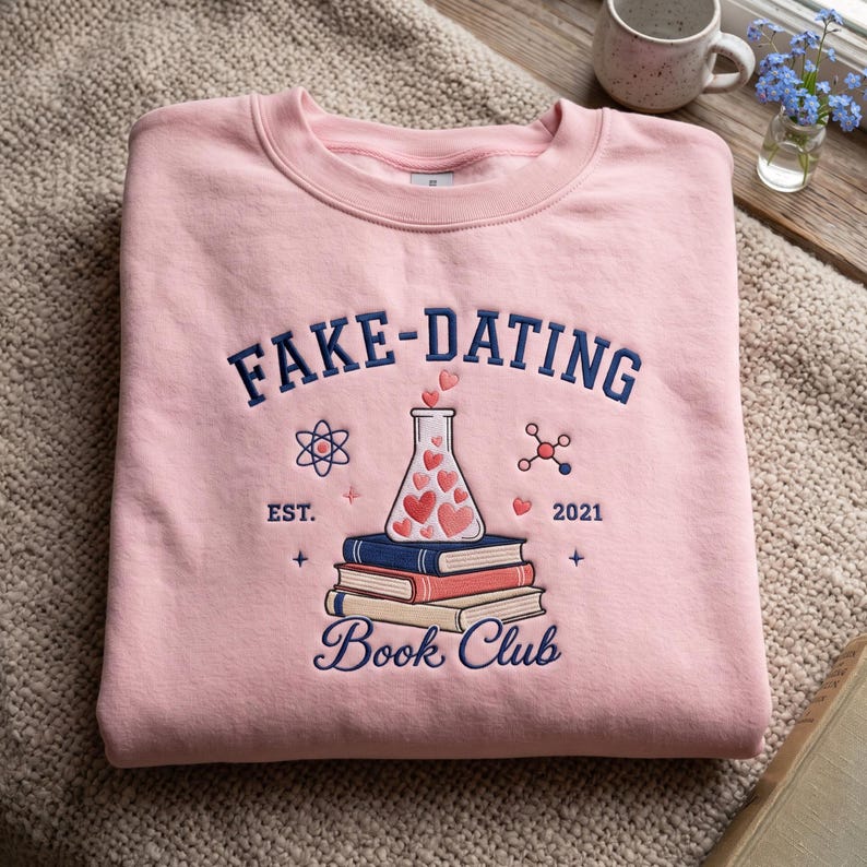 May include: A light pink crewneck sweatshirt featuring dark blue embroidered text that reads FAKE-DATING BOOK CLUB. The design includes a central Erlenmeyer flask filled with pink hearts, resting on a stack of three books, with science icons.