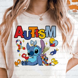 Op de afbeelding: Een wit T-shirt met een kleurrijke "AUTISM"-afbeelding met een blauw Stitch-personage uit Lilo & Stitch, omringd door puzzelstukjes, vogels en een hart.