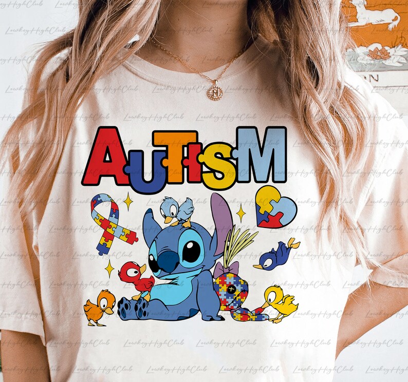Op de afbeelding: Een wit T-shirt met een kleurrijke "AUTISM"-afbeelding met een blauw Stitch-personage uit Lilo & Stitch, omringd door puzzelstukjes, vogels en een hart.