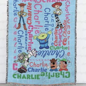 May include: A light blue woven throw blanket featuring the name Charlie repeated in various colors and fonts, alongside illustrations of Toy Story characters including Woody, Jessie, Buzz Lightyear, an alien, and two Mr. Potato Head figures.