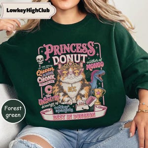May include: A forest green sweatshirt featuring a graphic of a calico cat wearing a crown and sunglasses, surrounded by text including Princess Donut, Queen Anne Chonk, and Best in Dungeon, along with a skull, a dinosaur, and a gold trophy.