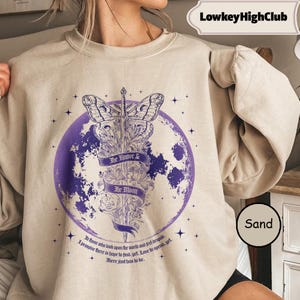 Könnte beinhalten: Ein sandfarbenes Sweatshirt mit Rundhalsausschnitt und einer violetten Grafik eines Nachtfalters auf einem Schwert, zentriert in einem Vollmond. Das Design enthält florale Elemente, Sterne und ein Banner mit dem Text The Knave & The Moon.