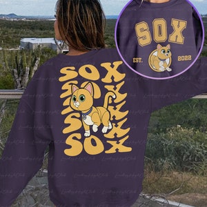 May include: Purple sweatshirt with a yellow and orange graphic of a cat with the word "SOX" repeated multiple times. The cat is wearing a collar with a tag. The text "EST. 2022" is also on the sweatshirt.